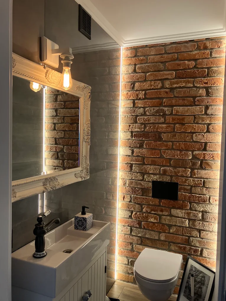 Loft w małej skali — Ceglana ściana i ciepłe światło w niewielkiej łazience. Jasna ceramika równoważy mocną fakturę.